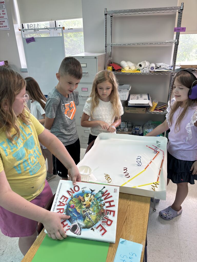 Students gather around a tabletop marble run built with straws and pipe cleaners, exploring motion and design through STEM learning.