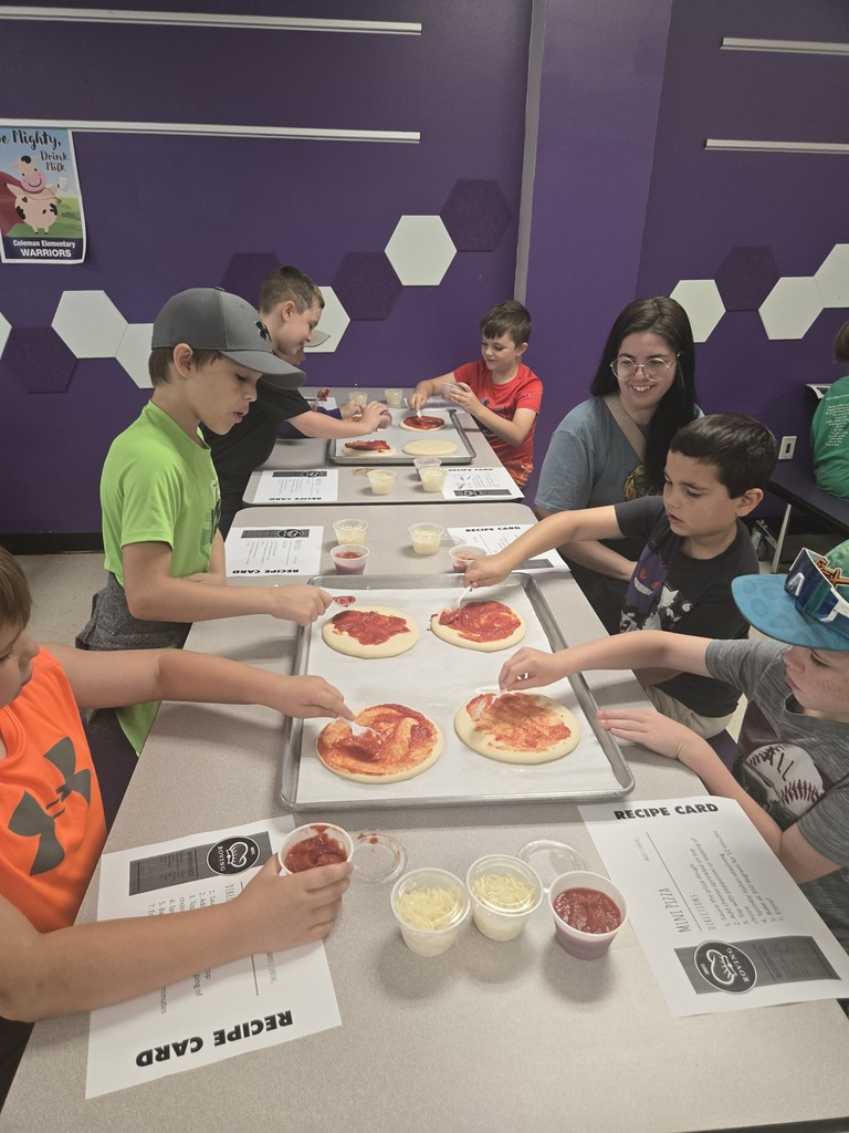 Students spread pizza sauce onto personal pizzas during a hands-on cooking activity, following recipe cards and working together at tables.