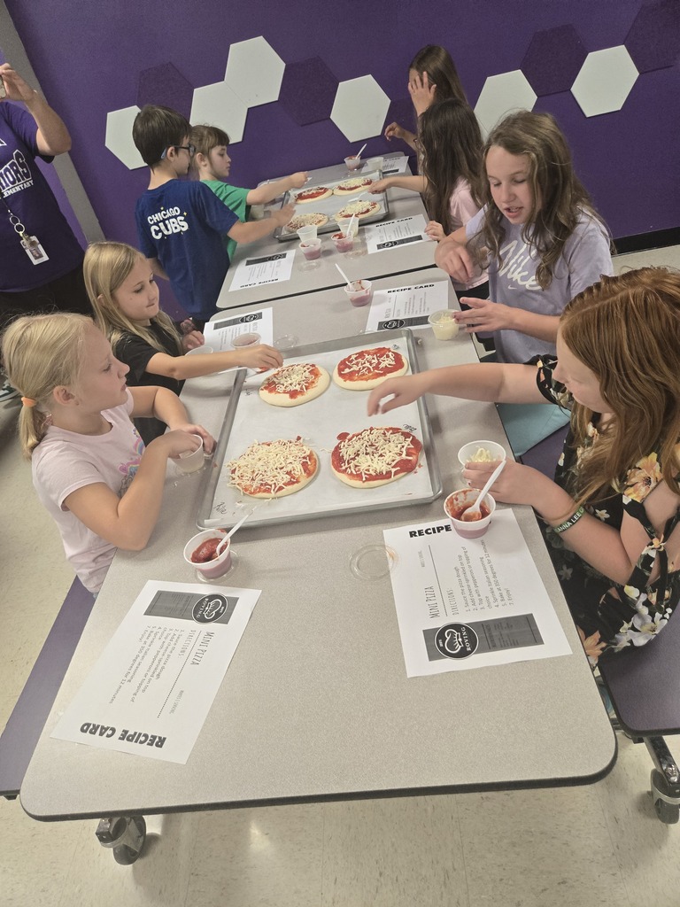 Students top personal pizzas with cheese and pepperoni during a classroom cooking lesson, using recipe cards to guide the process.