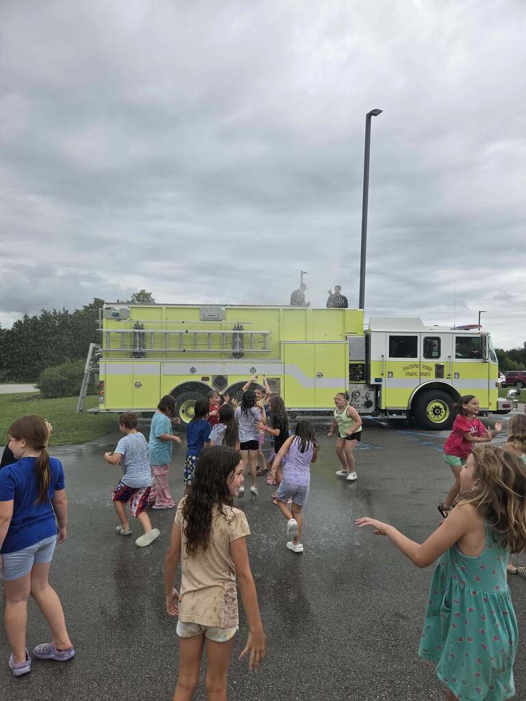 Students run and play in water sprayed from a fire truck during an outdoor school field day activity.