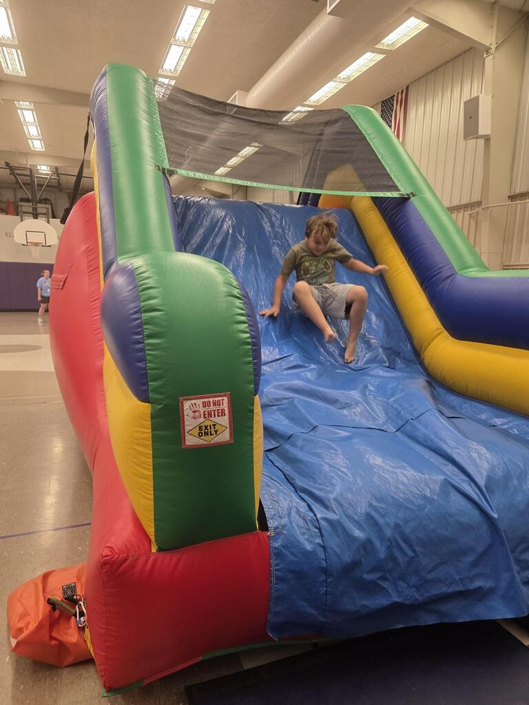 Student slides down a colorful inflatable slide during an indoor school celebration in the gymnasium.