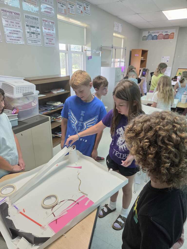 Students test a handmade marble run in a classroom, watching a ball travel through ramps, tubes, and obstacles.