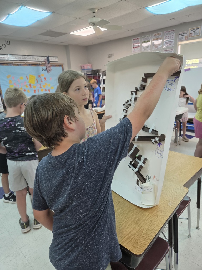 Students demonstrate a vertical cardboard marble run, guiding a ball through winding tracks and interactive sections.