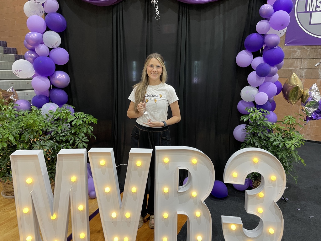 Staff member poses with an award behind illuminated “MVR-III” letters, framed by purple balloons and decorative plants at a recognition event.