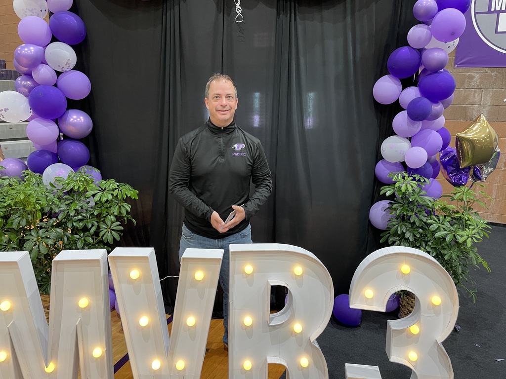 Award recipient stands in front of illuminated MVR-III letters and a purple balloon display, holding a recognition award at a school celebration.