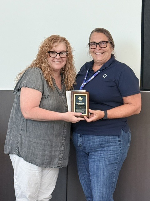 Two women smile while holding a recognition plaque together during an award presentation.