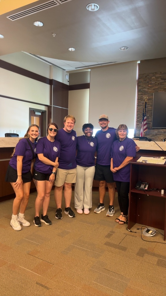 Six staff members wearing matching purple school shirts pose together inside a meeting room.