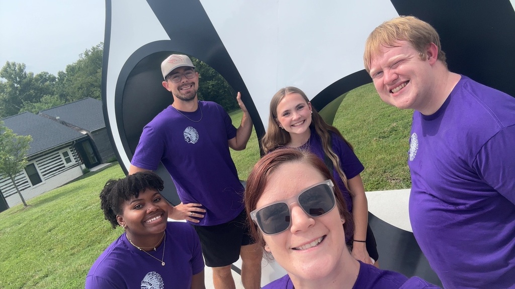 Five smiling staff members in matching purple school shirts pose for a selfie outdoors beside a large sculpture.