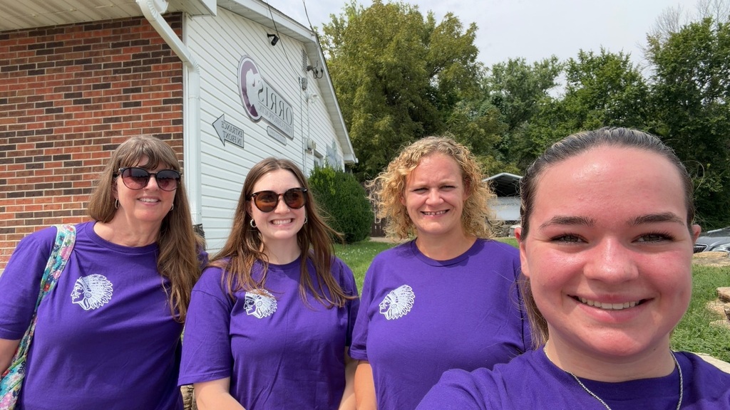 Four smiling staff members in matching purple school shirts take a selfie outside a community building.