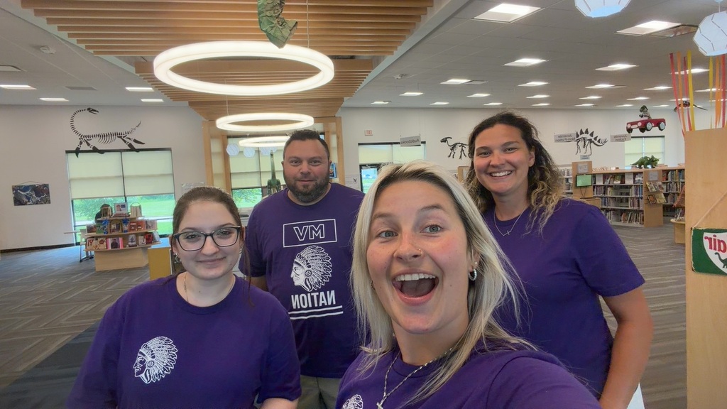 Four smiling staff members in purple school shirts take a selfie inside a bright library.