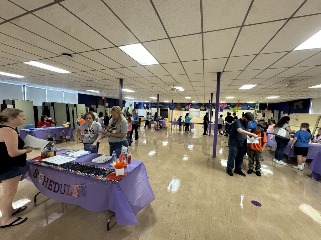 Families and staff visit information and scheduling tables during a school event in a large multipurpose room.