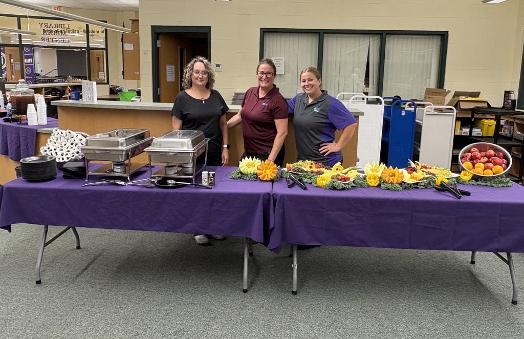 Three smiling staff members stand behind a buffet table with serving trays and colorful fruit and vegetable platters.