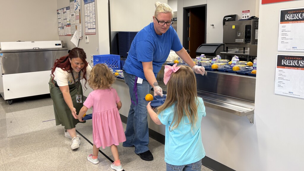 Two staff members handing two young students trays of food.