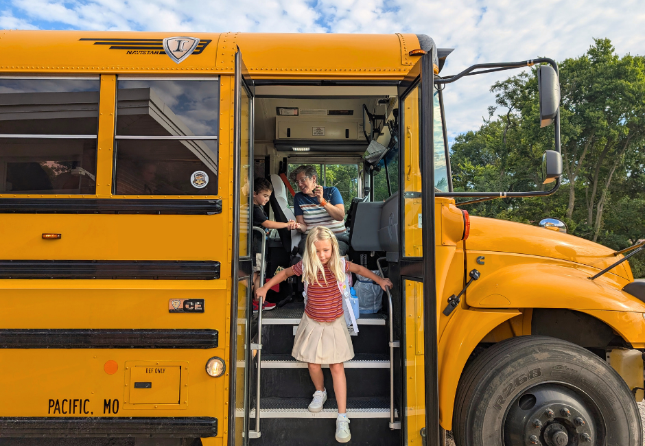 A young student steps carefully off a yellow school bus while an adult and another student wait behind her.