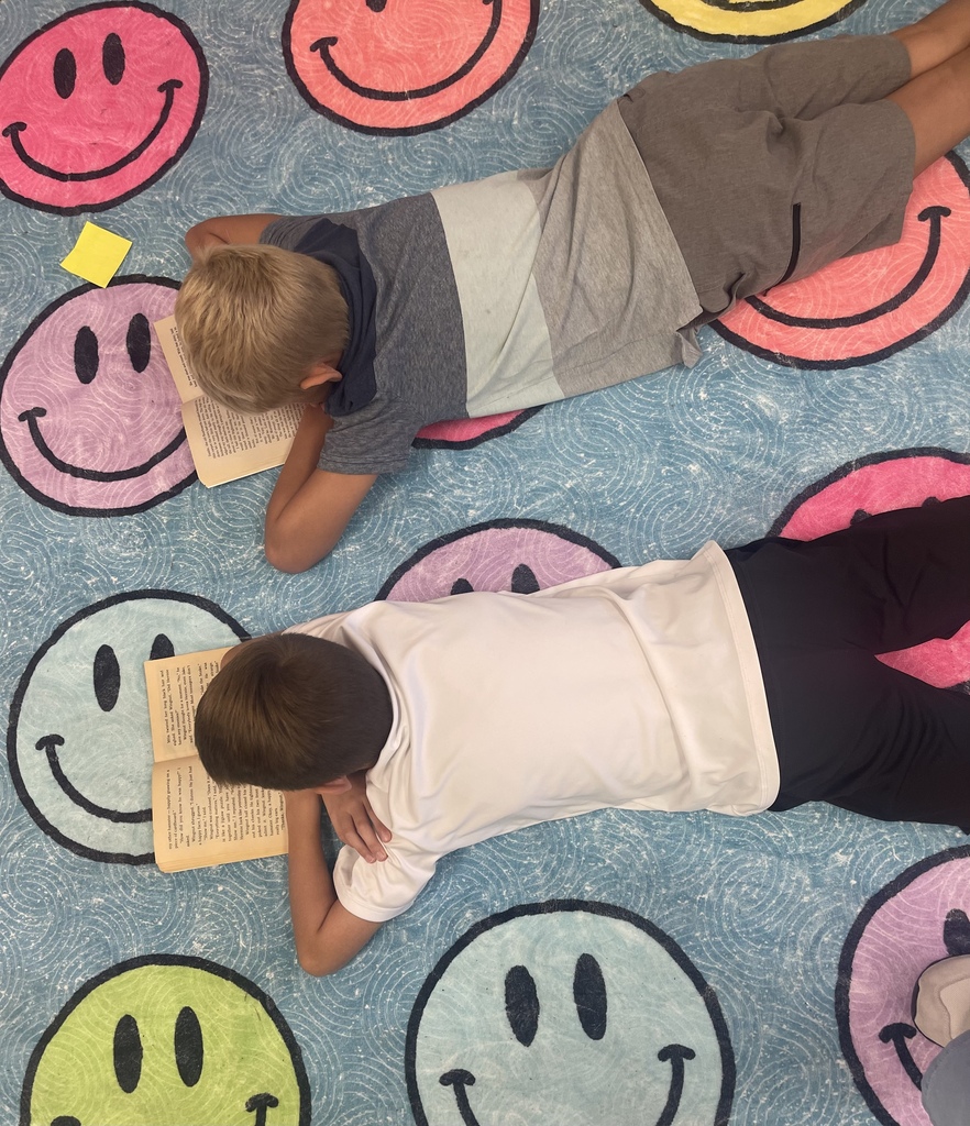Two students laying on a smiley face rug while reading a book.