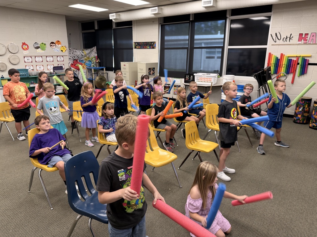 A group of students playing with pool noodles in a music classroom.