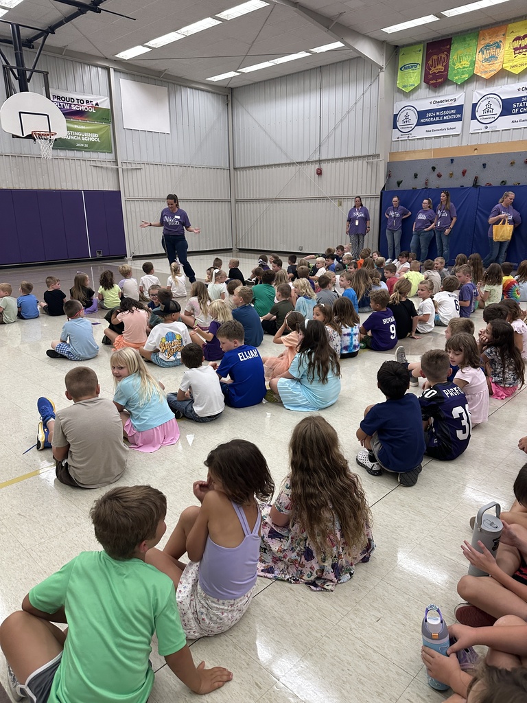 Elementary students sit together on a gym floor while a teacher addresses the group, with several staff members nearby.