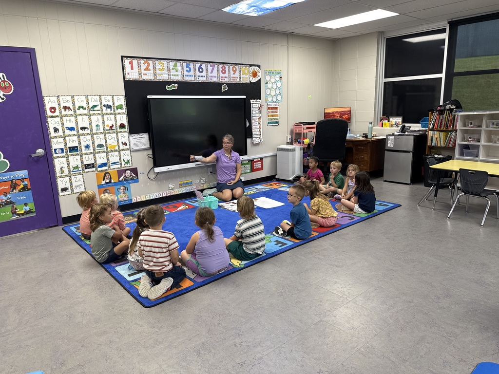 Two elementary classrooms show teachers leading groups of young students seated together on classroom rugs.