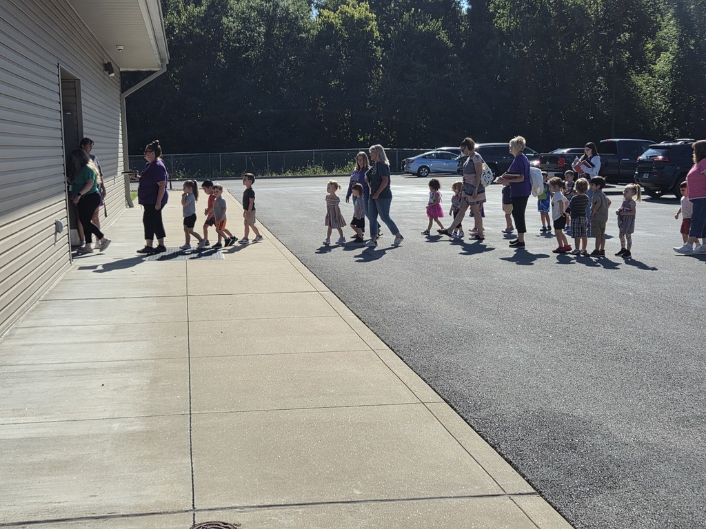Young children and adults walk in a line across a sunny parking lot toward the entrance of a school building.