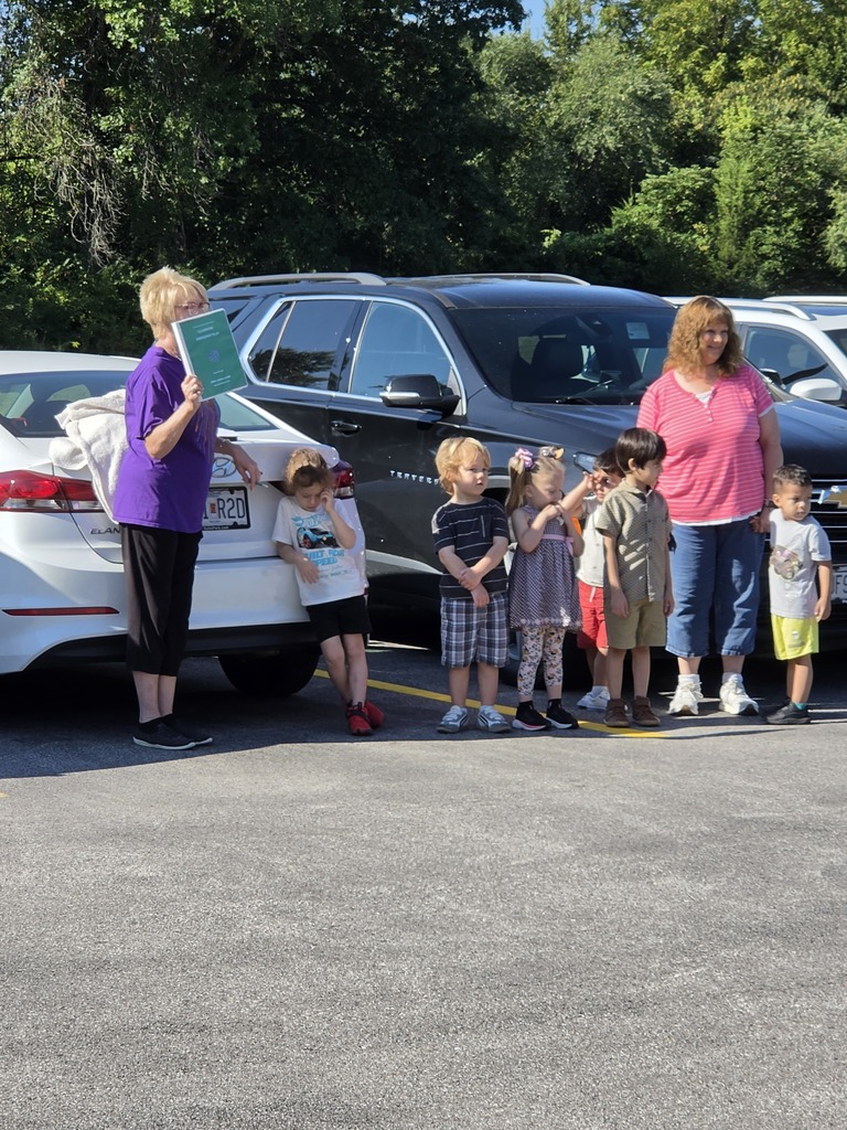 Young children stand with two adults in a sunny parking lot, preparing to walk together toward a building.