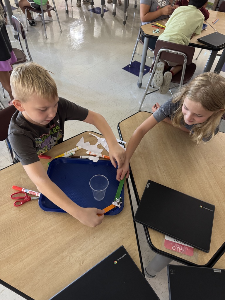 Two students work together on a classroom challenge using craft sticks, paper, and a plastic cup.