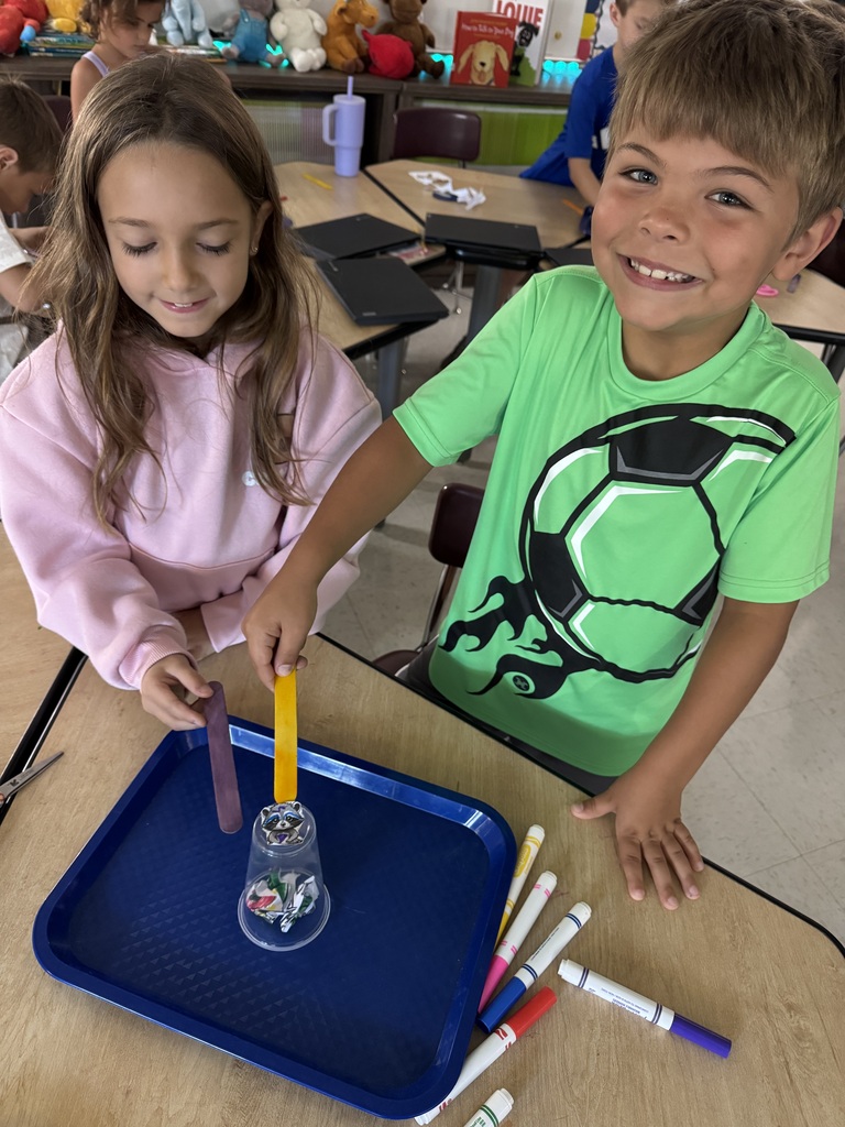 Two smiling students test a structure made from a plastic cup, paper, and colorful craft sticks.
