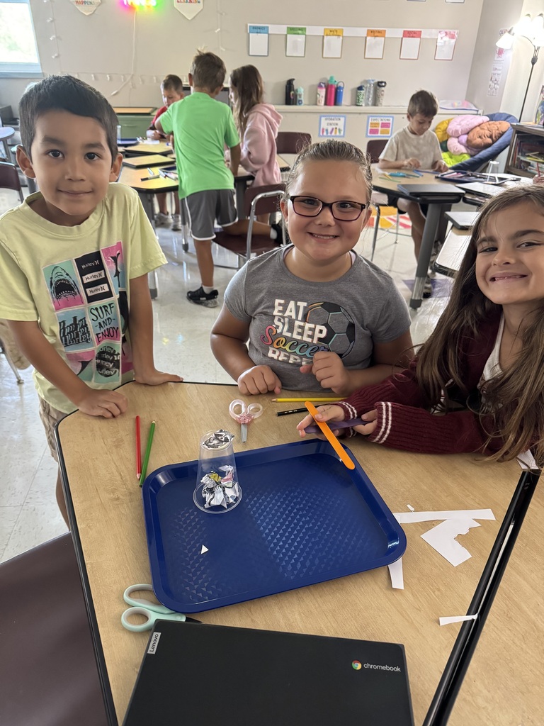 Three students pose beside their completed classroom project, surrounded by craft supplies and a Chromebook.