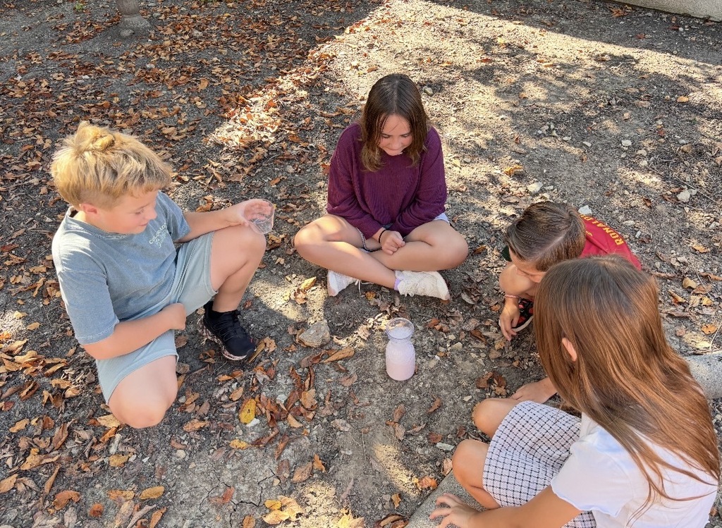Four students sit in a circle outdoors, watching a pink mixture foam from a container during a science experiment.