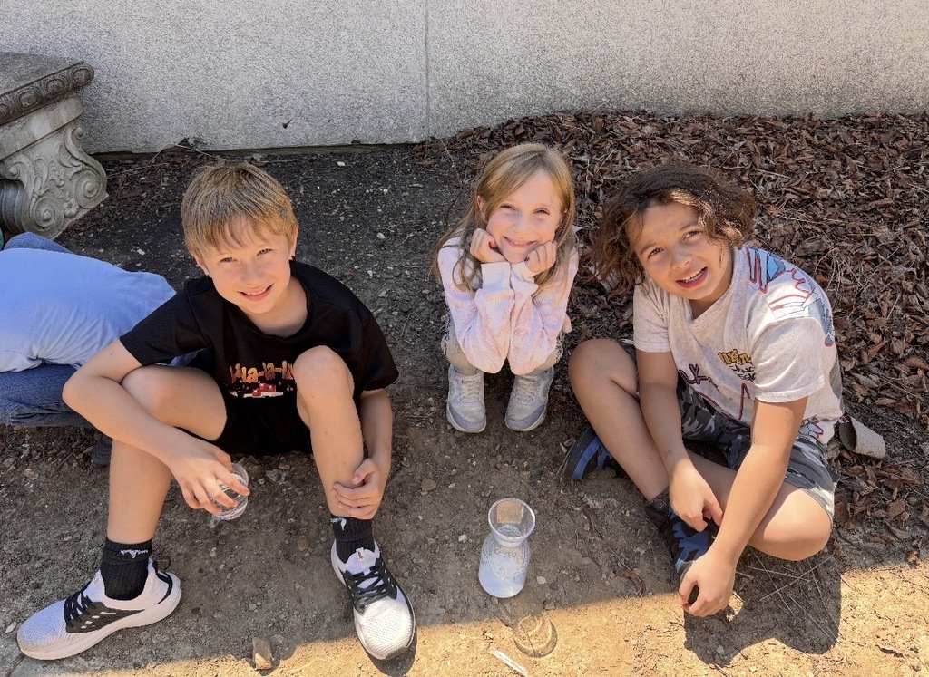 Three students sit outdoors around a container, smiling as they observe a foamy science experiment.