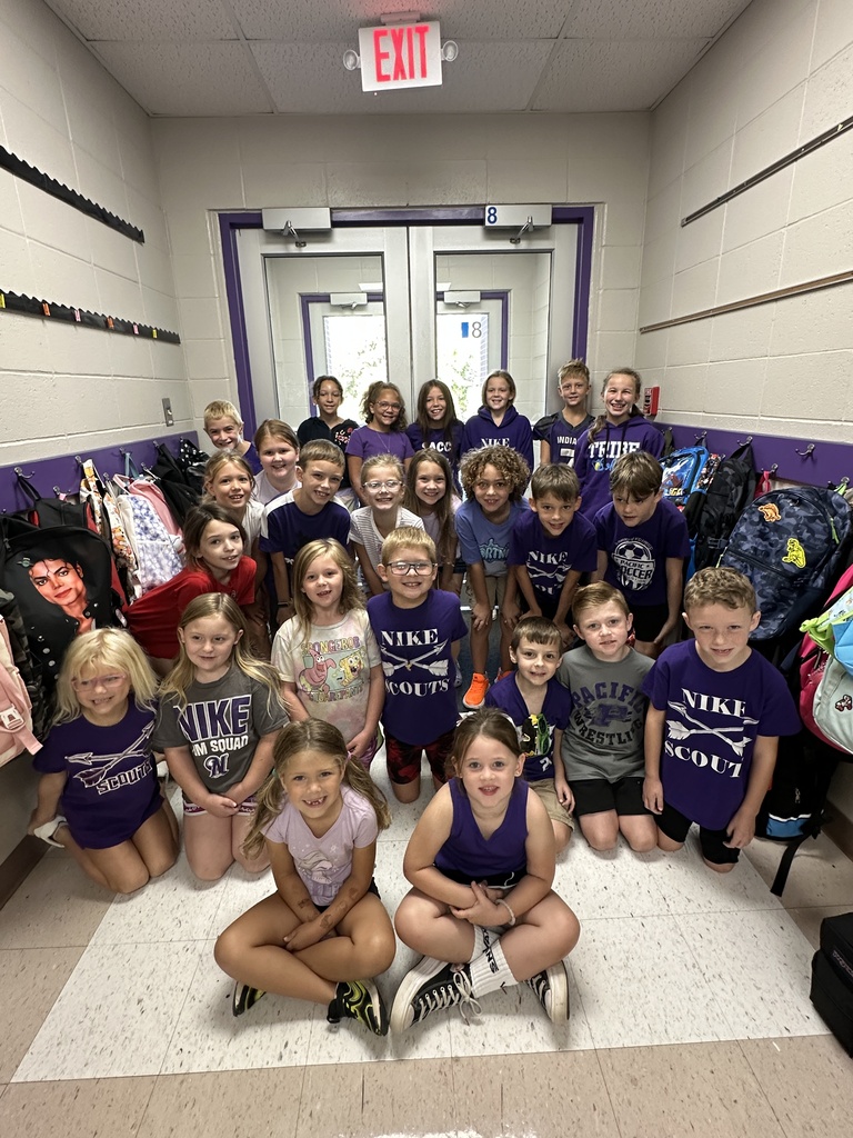 A large group of elementary students, many dressed in purple, smile together for a group photo in a school hallway.