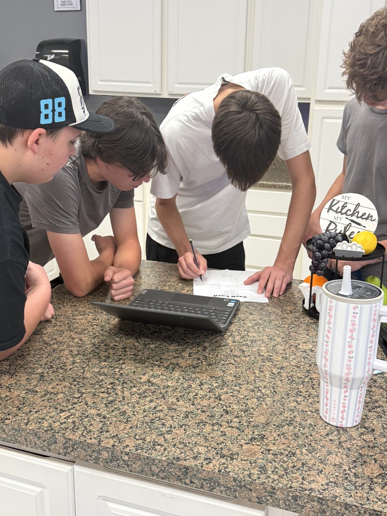 Four students collaborate at a classroom counter as one writes on a worksheet beside an open laptop.