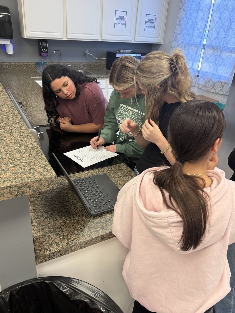 Four students gather around a classroom counter, working together on a worksheet beside an open laptop.