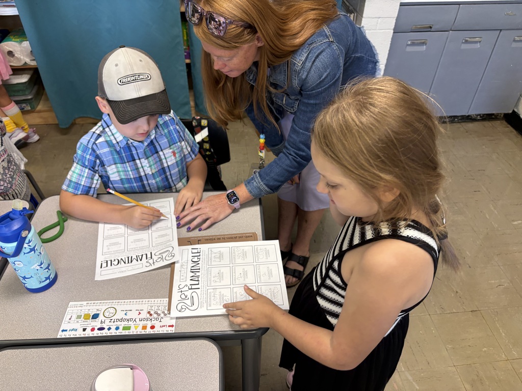 A teacher leans over a classroom table to assist two students completing โLetโs Flamingleโ worksheets.