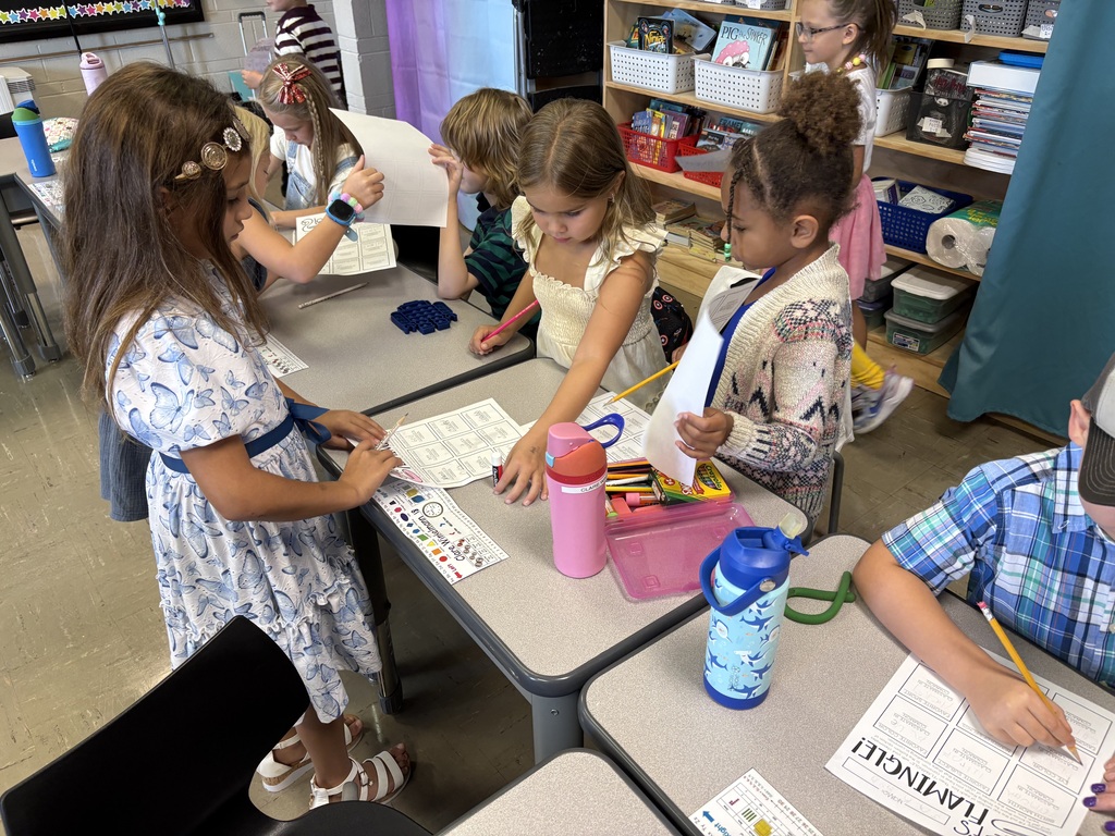 Students move around classroom tables, writing on worksheets and interacting with classmates during an activity.
