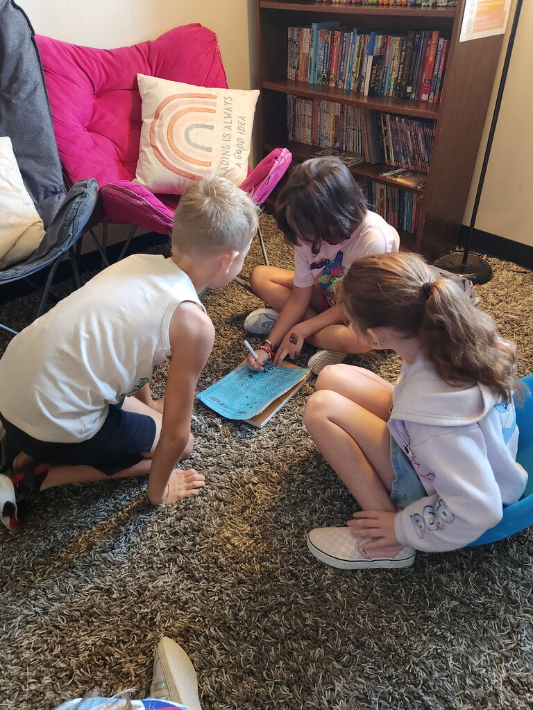 Three students sit together on a classroom rug, collaborating on a worksheet near a bookshelf and reading chair.