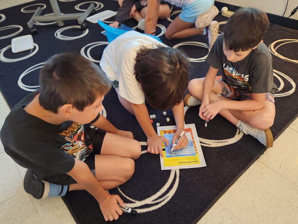Three students sit on a classroom rug, using dice and markers to complete a small-group learning activity.