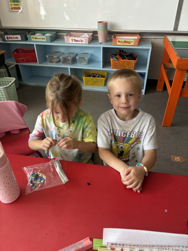 Two students sit at a classroom table making colorful beaded bracelets together.