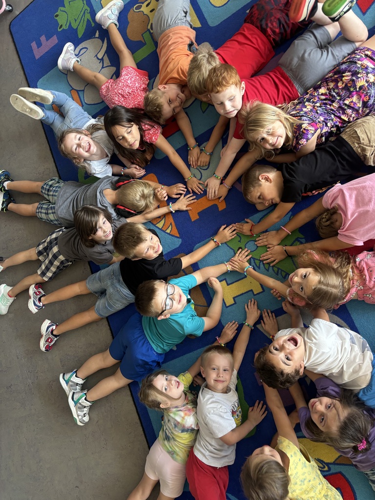 Students lie in a circle on a classroom rug, reaching toward the center to display their colorful bracelets.
