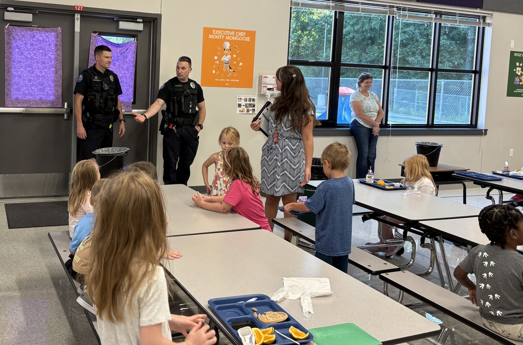 Two uniformed police officers speak with students and staff in a school cafeteria.