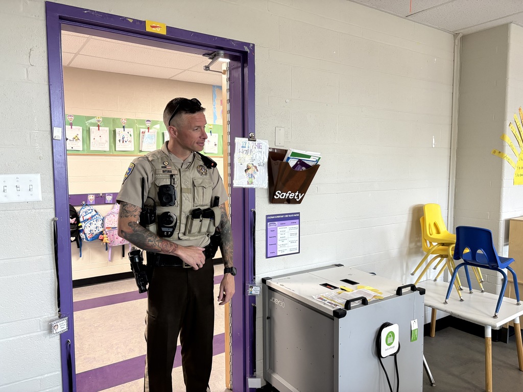 A uniformed law enforcement officer stands in a classroom doorway during a school visit.