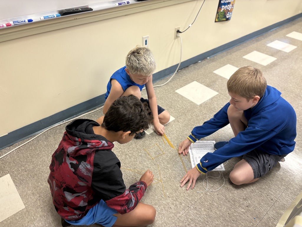 Three students sit on a classroom floor, working together to build geometric shapes with thin sticks.