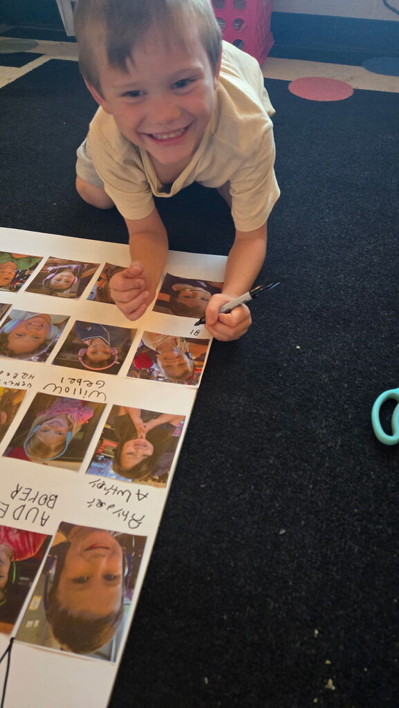 Smiling student kneels on a classroom rug, writing on a poster filled with classmates’ photos and names.