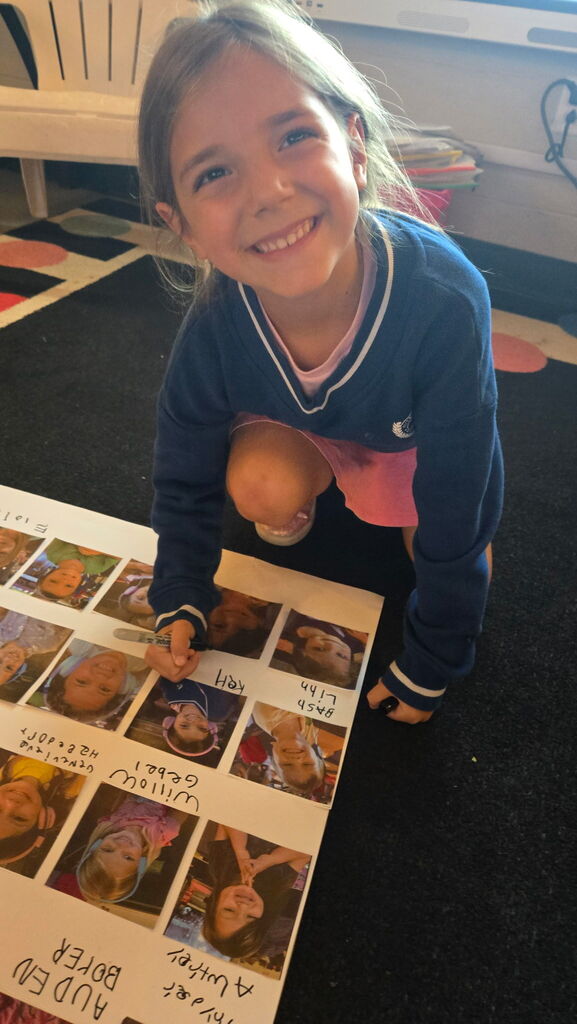 Smiling student kneels beside a class photo poster, adding a handwritten name with a marker.