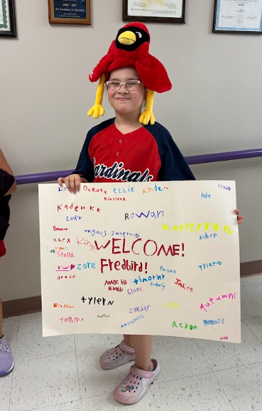 Student wearing a plush cardinal hat holds a colorful handmade sign reading โWELCOME! Fredbird!โ