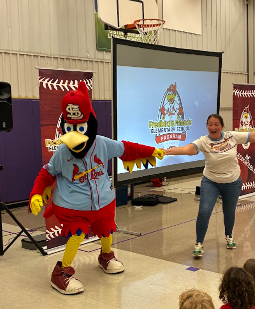 Fredbird holds hands with a smiling presenter during a lively elementary school assembly.