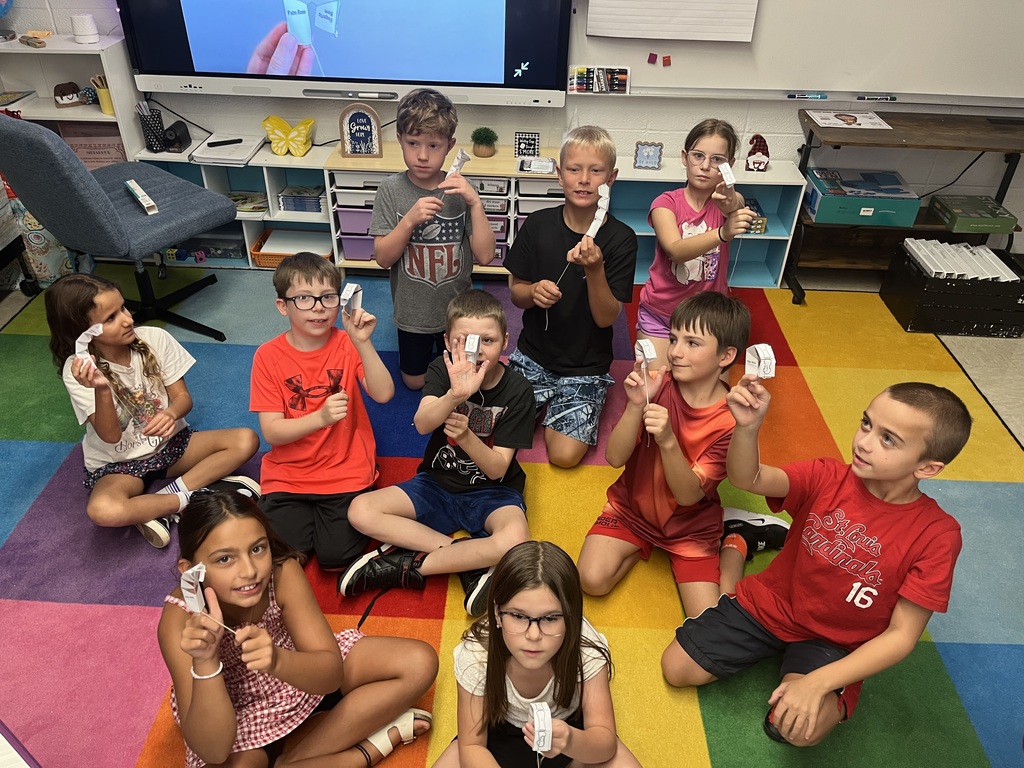 Students sit together on a colorful classroom rug, proudly holding up their completed paper-and-string creations.