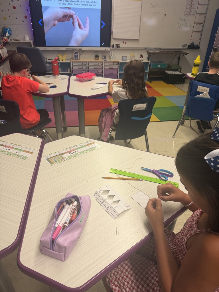 Students follow classroom instructions to fold paper and attach string while creating small paper models at their desks.