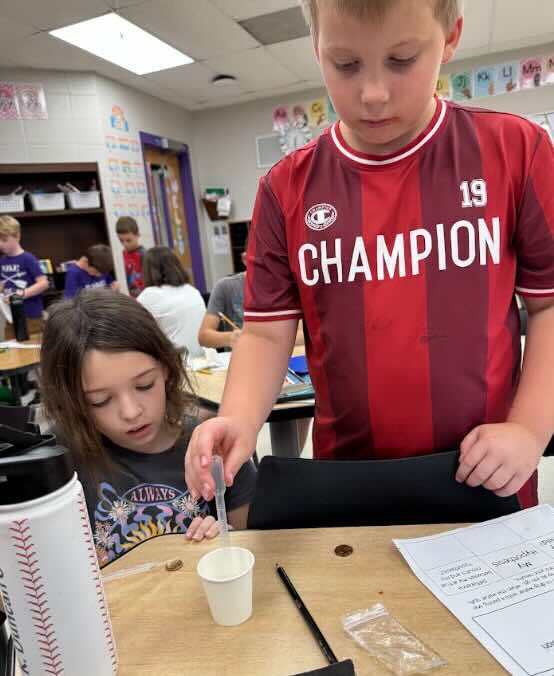 Two students work together using a dropper and cup while conducting a penny and water surface tension experiment.