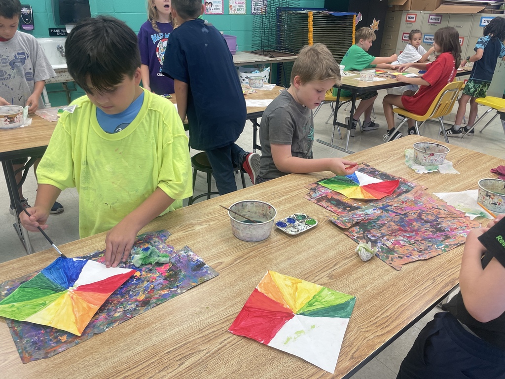 Students paint bright color-wheel designs at classroom tables during a hands-on art lesson.