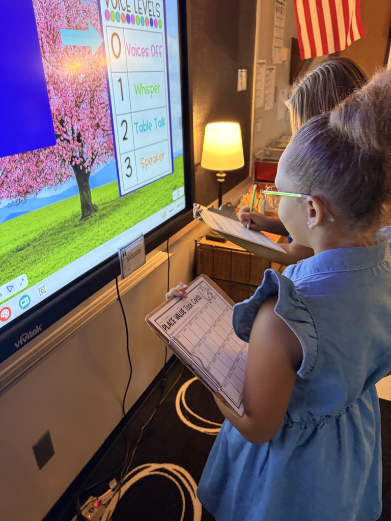 Students complete place-value task cards on clipboards beside a classroom display showing voice-level expectations.